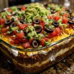 Close-up view of a fully loaded Seven layer dip in a clear glass baking dish, topped with guacamole, tomatoes, olives, and cheese.