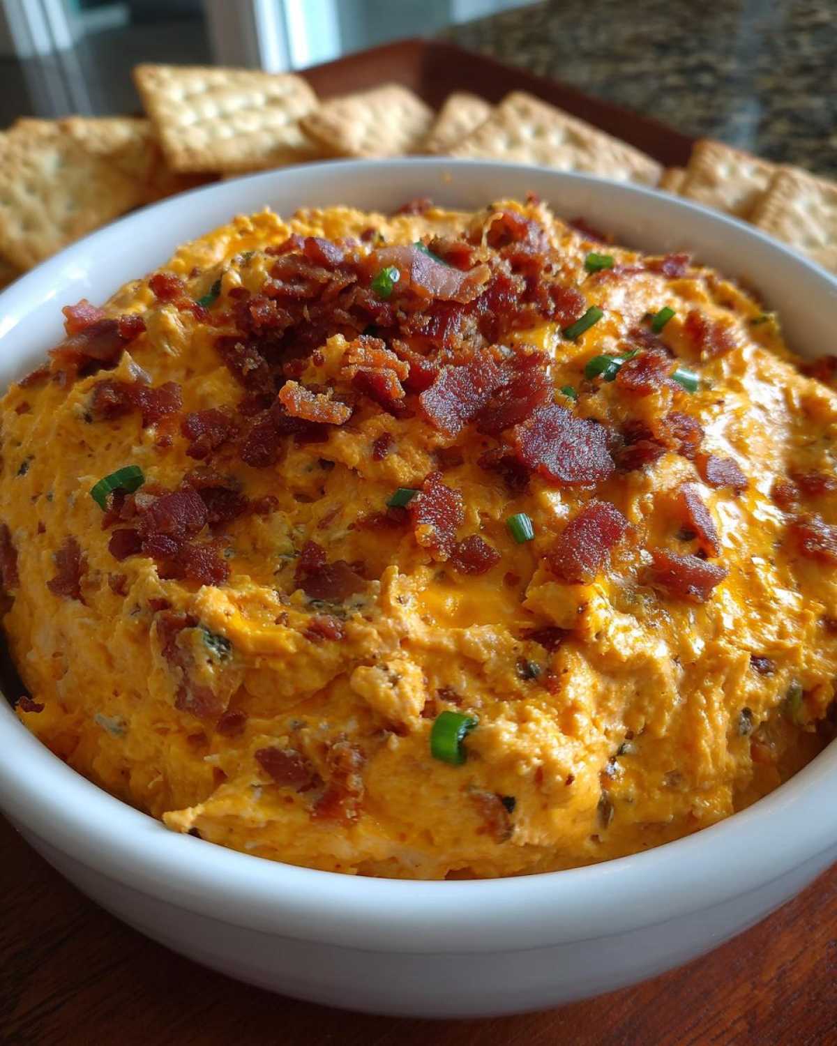A close-up of creamy, orange-hued million dollar dip topped generously with crumbled bacon and fresh chives.