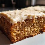 Close-up of a moist slice of applesauce cake topped with thick, creamy frosting dusted with spice.
