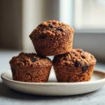 Three dark, moist bran muffins studded with raisins, stacked on a small white plate.