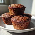 Four dark, moist bran muffins stacked on a white plate, highlighted by strong sunlight.