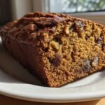 Close-up of a moist slice of dark brown date nut bread showing visible dates and walnuts, resting on a white plate.