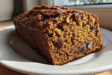 Close-up of a moist slice of dark brown date nut bread showing visible dates and walnuts, resting on a white plate.