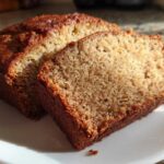 Close-up of a moist slice of sugar free banana bread next to the rest of the loaf on a white plate.