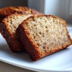 Two moist slices of sugar free banana bread resting on a white plate, showing a rich brown crust.