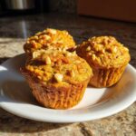 Three freshly baked morning glory muffins topped with chopped walnuts served on a white plate.