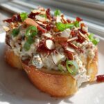 A thick layer of creamy Neiman Marcus dip topped with slivered almonds, bacon bits, and green onions on toasted bread.