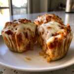 A close-up of a pizza muffin cut in half, showing the soft interior topped with melted mozzarella cheese and tomato sauce, part of a batch of pizza muffins.