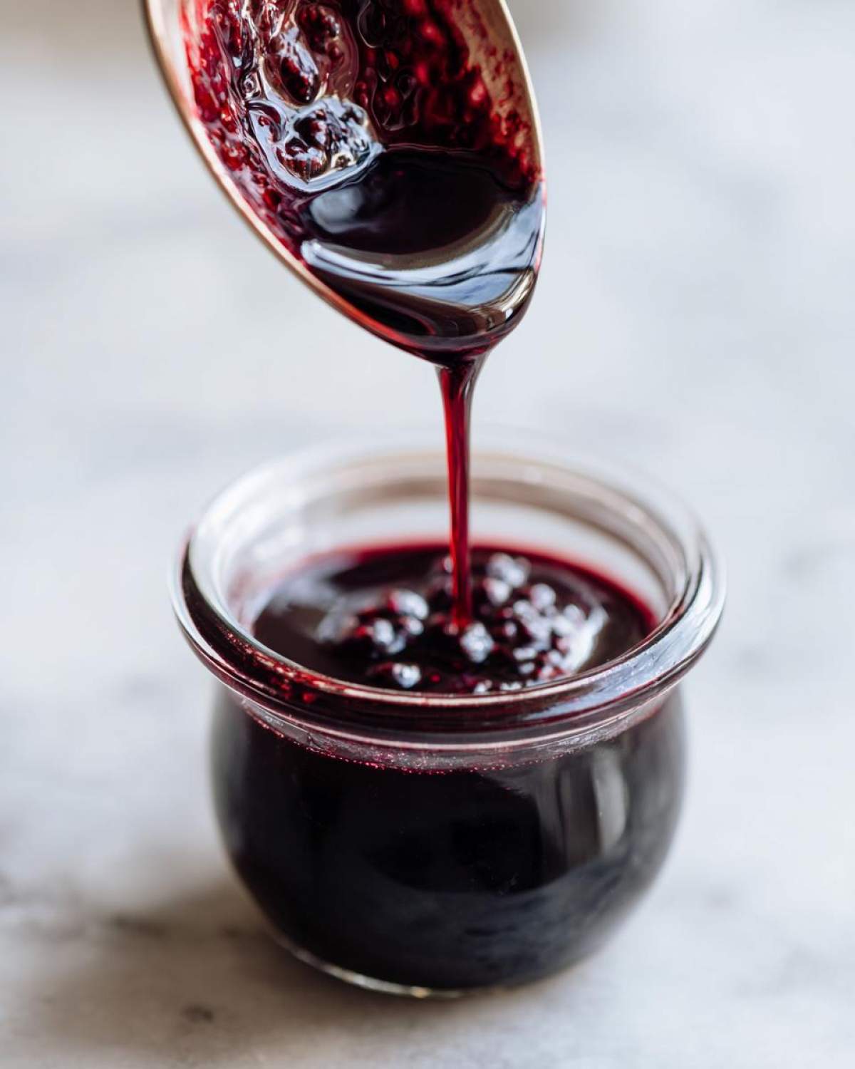 Close-up of thick, dark purple elderberry syrup being poured from a spoon into a small glass jar.