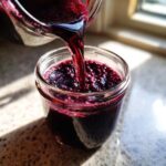 Close-up of rich, dark purple elderberry syrup being poured from a pitcher into a small glass jar.