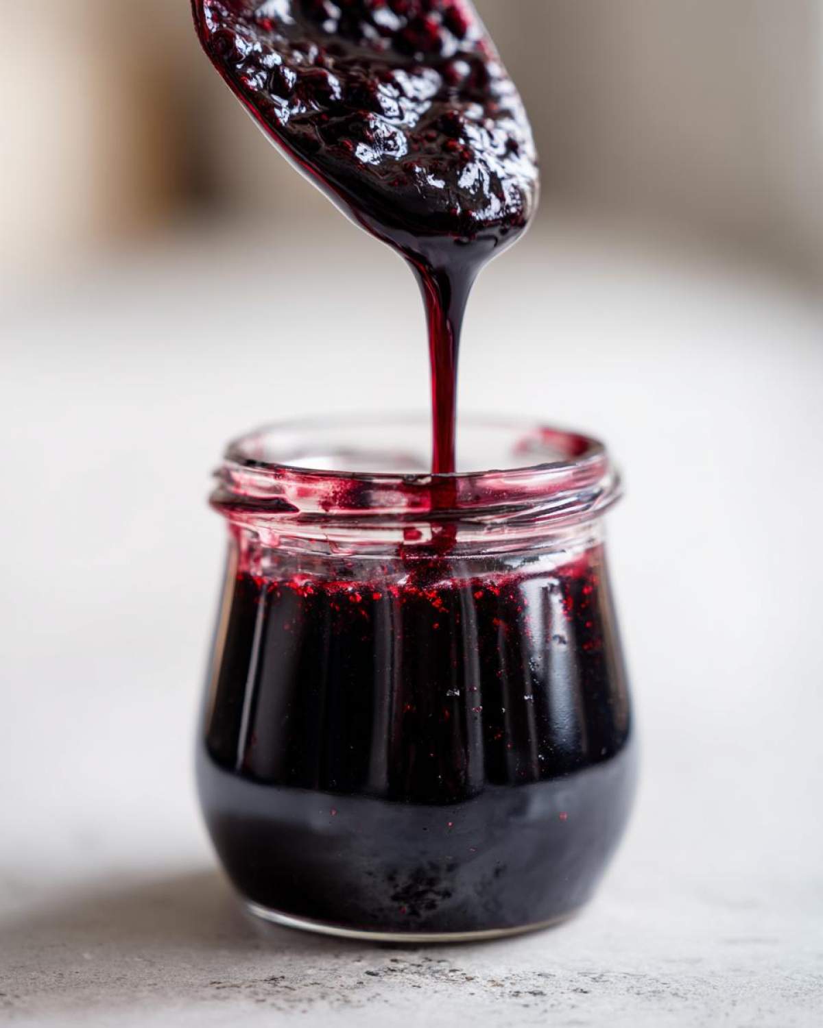 Close-up of thick, dark elderberry syrup being poured from a spoon into a small glass jar.