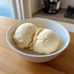 Two scoops of creamy, pale yellow protein ice cream served in a white bowl on a wooden table.