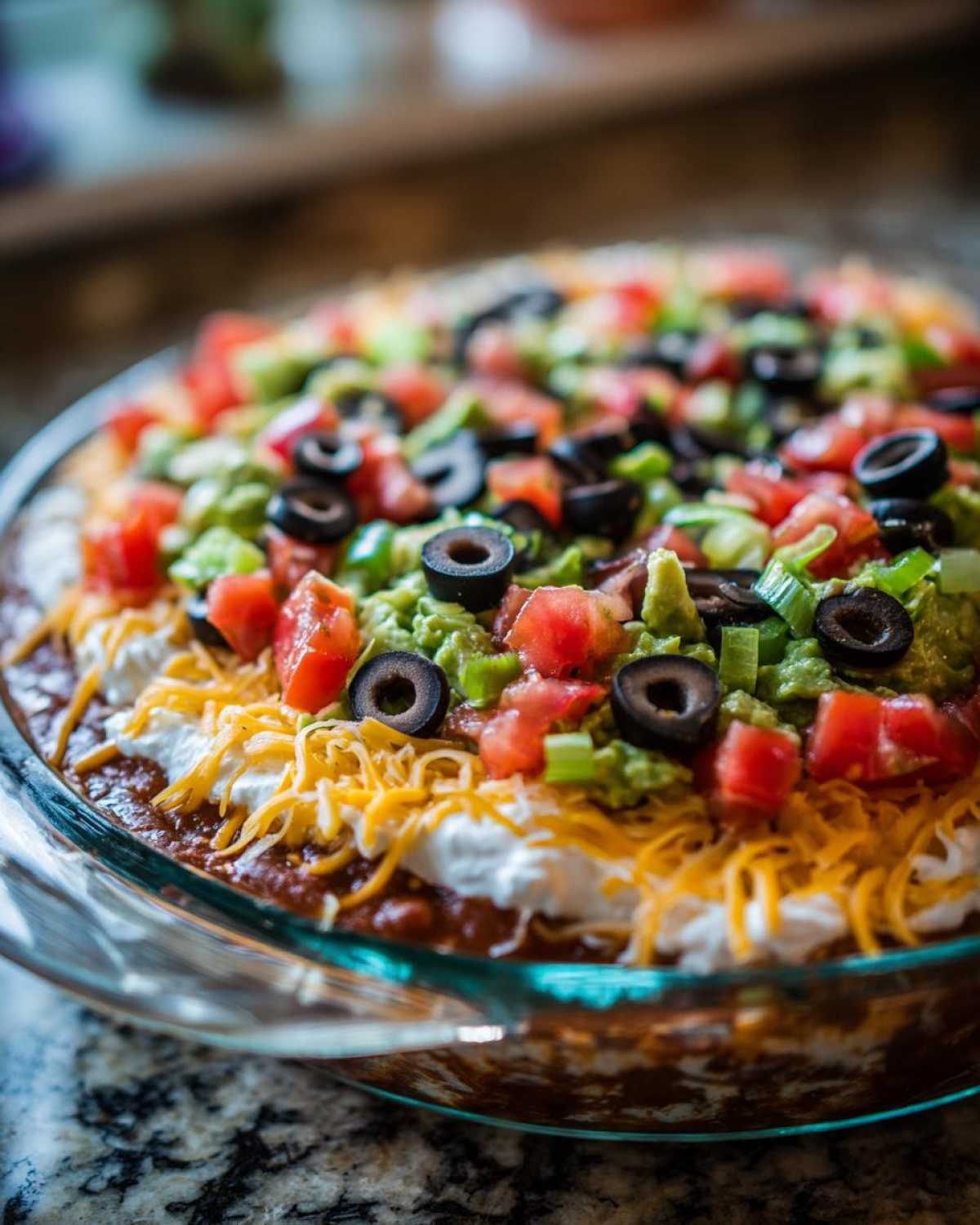 Close-up of a vibrant Seven layer dip in a clear glass dish, topped with guacamole, tomatoes, olives, and shredded cheese.