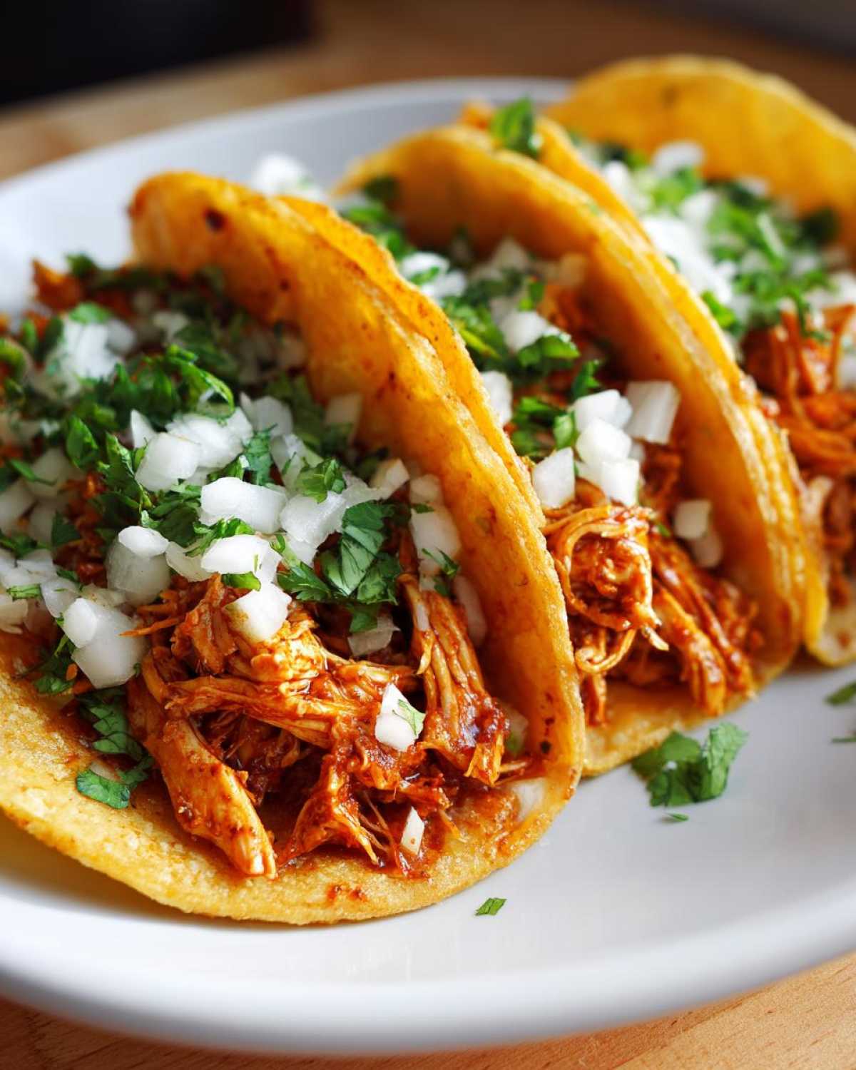 Three appetizing chicken tacos filled with shredded, saucy chicken, topped with diced white onion and fresh cilantro.