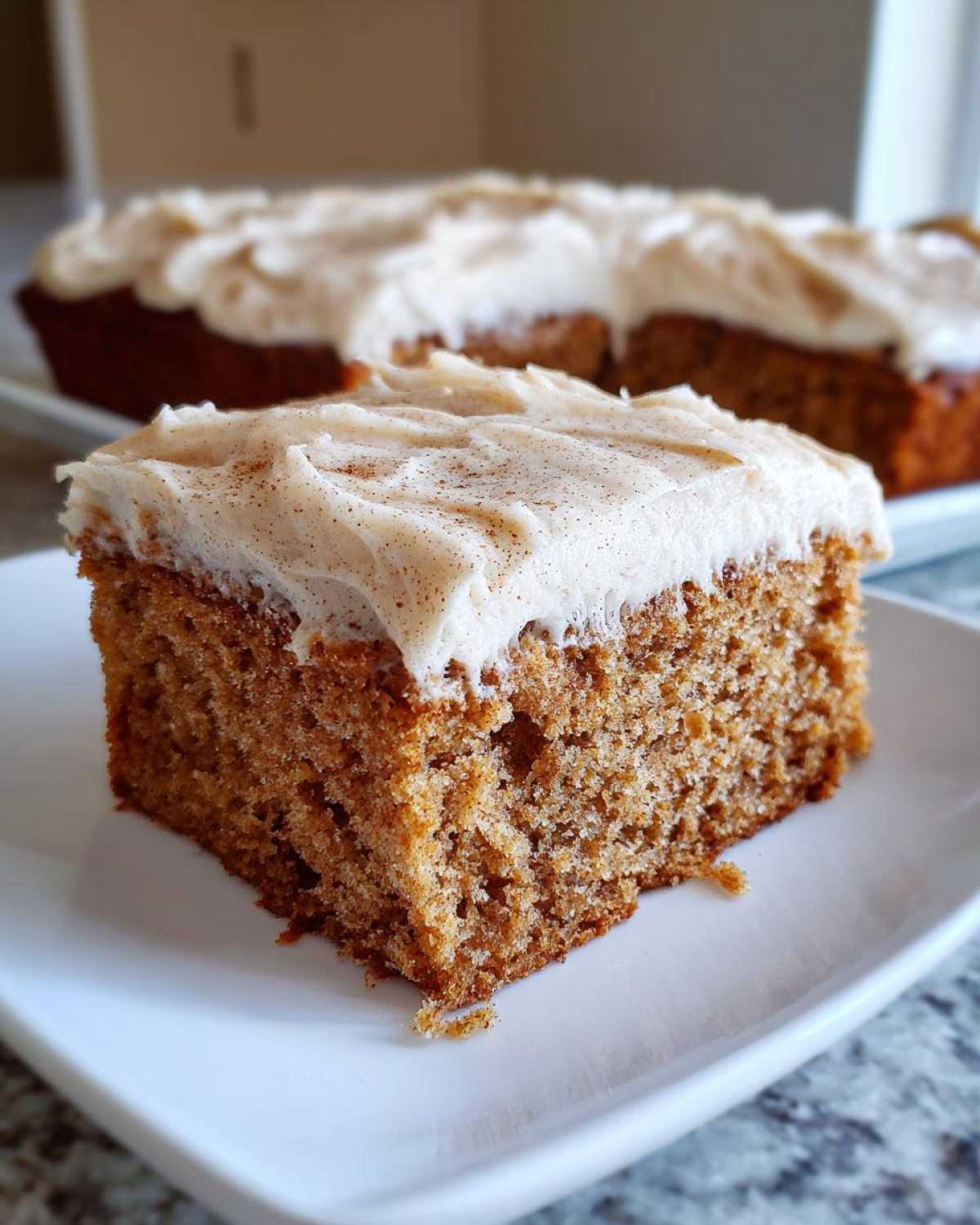 A close-up of a moist slice of applesauce cake topped with thick, creamy frosting dusted with cinnamon.