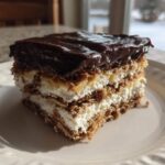 A decadent slice of eclair cake featuring layers of graham crackers, cream filling, and topped with shiny chocolate ganache.