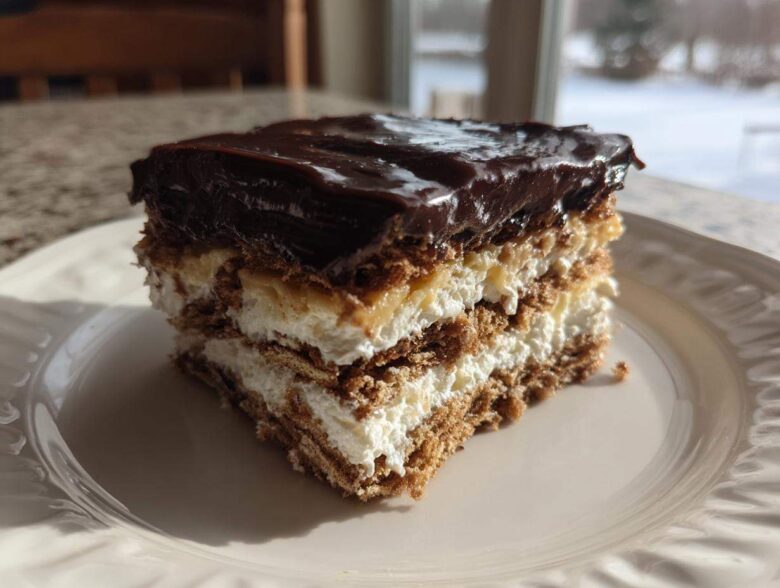 A decadent slice of eclair cake featuring layers of graham crackers, cream filling, and topped with shiny chocolate ganache.