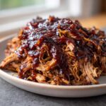 Close-up of tender Slow cooker pulled pork generously topped with rich, dark BBQ sauce on a light plate.