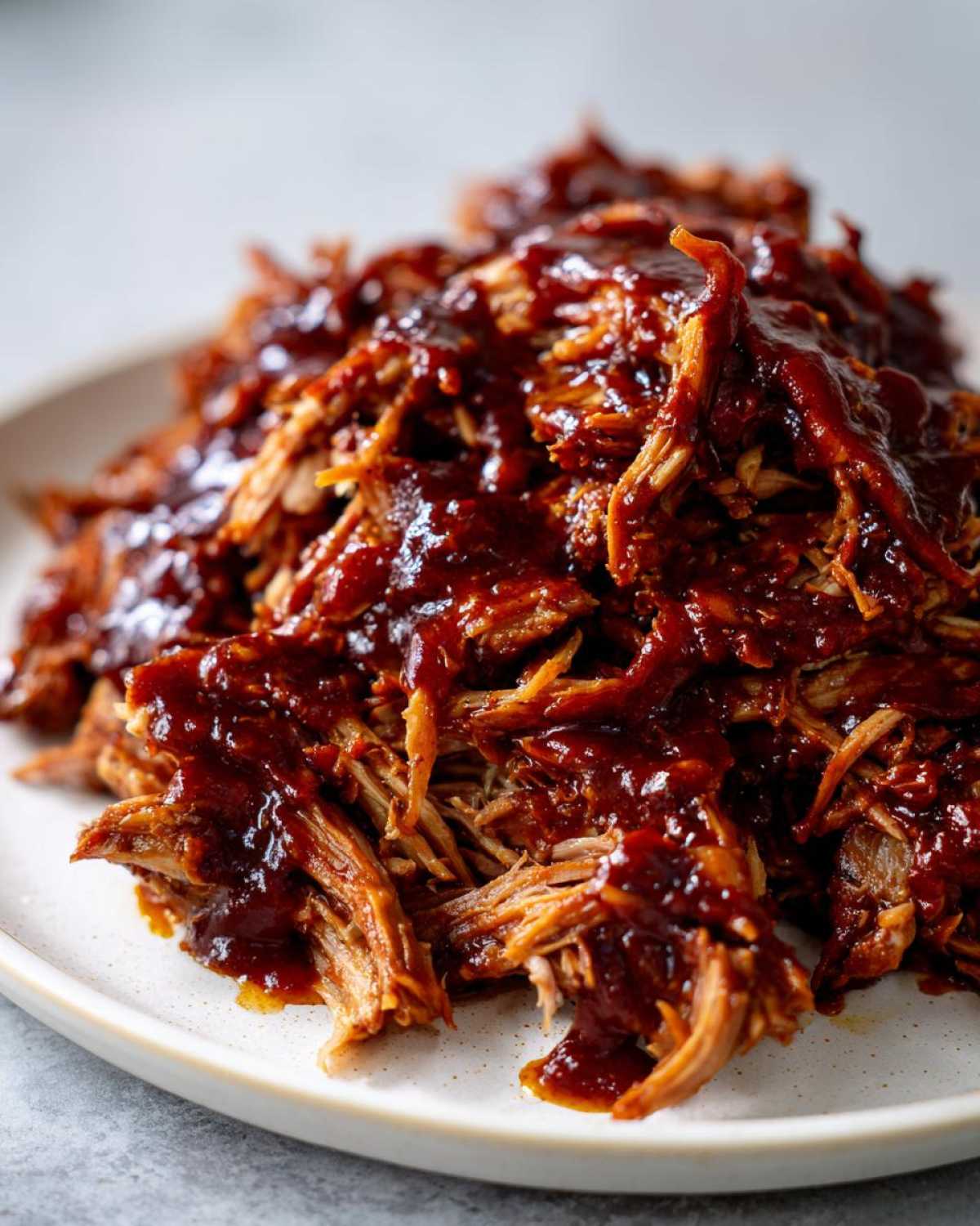 Close-up of tender, shredded Slow cooker pulled pork heavily coated in rich, dark barbecue sauce on a white plate.