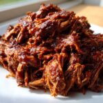A close-up of shredded, saucy Slow cooker pulled pork piled high on a white plate, glistening under natural light.