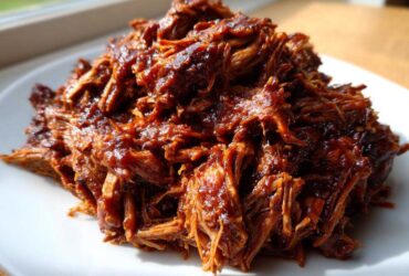 A close-up of shredded, saucy Slow cooker pulled pork piled high on a white plate, glistening under natural light.
