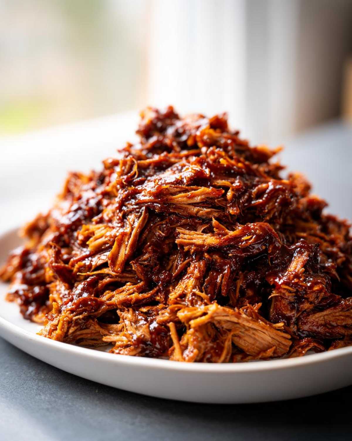 A close-up of a mound of saucy, shredded Slow cooker pulled pork piled high on a white plate.