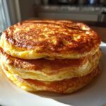 A close-up shot of three very thick and fluffy pancakes stacked high on a white plate, perfectly golden brown.