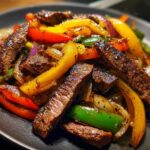 Close-up of perfectly cooked steak fajitas with colorful bell peppers and onions on a dark plate.