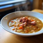 A steaming white bowl filled with thick Ham and Bean Soup, topped with diced ham pieces.