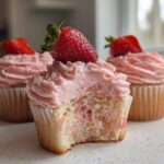 Close-up of a delicious strawberry cupcake with pink frosting and a fresh strawberry on top; a bite is missing to show the cake texture.