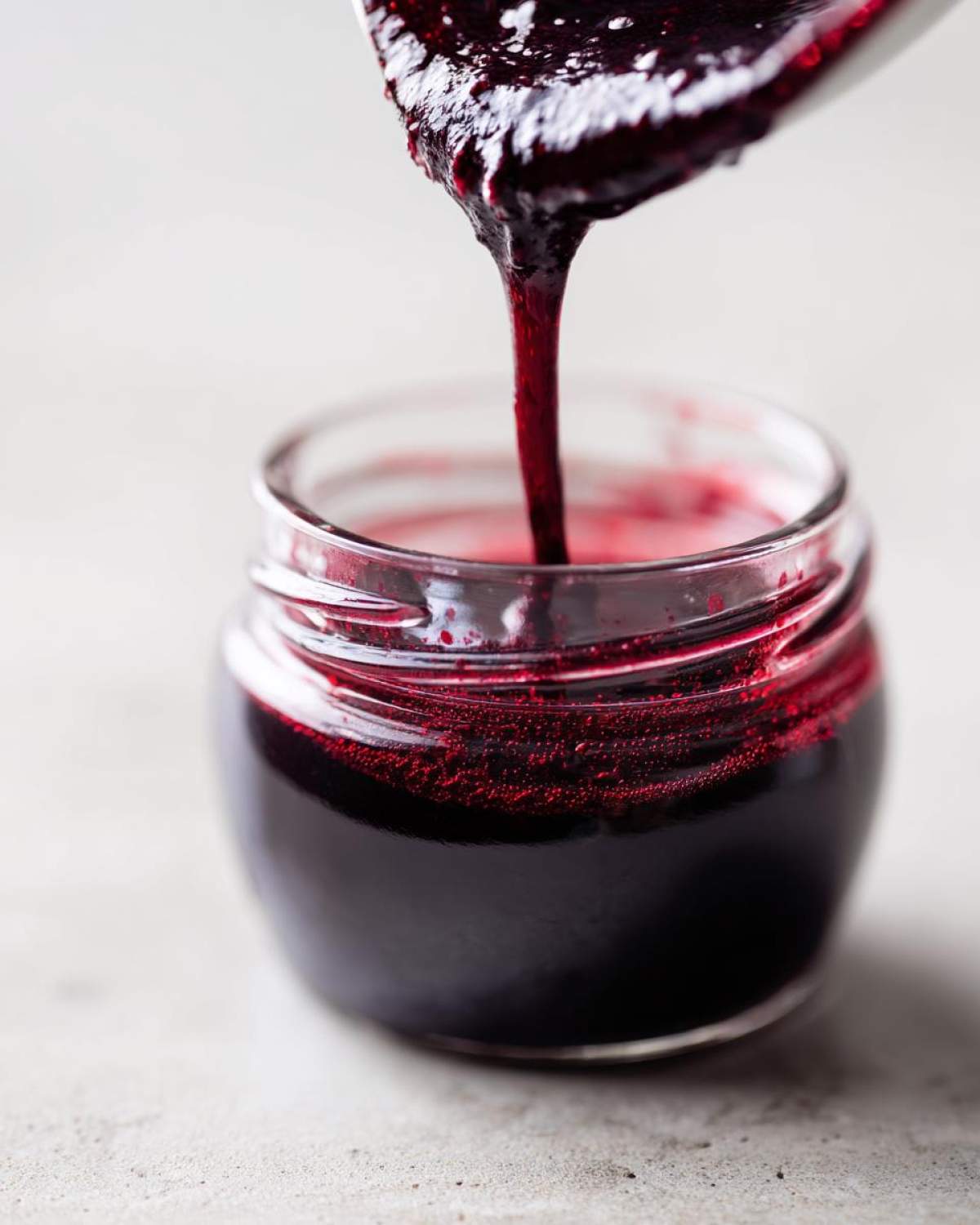 Close-up of thick, dark purple elderberry syrup being poured from a spoon into a small glass jar.