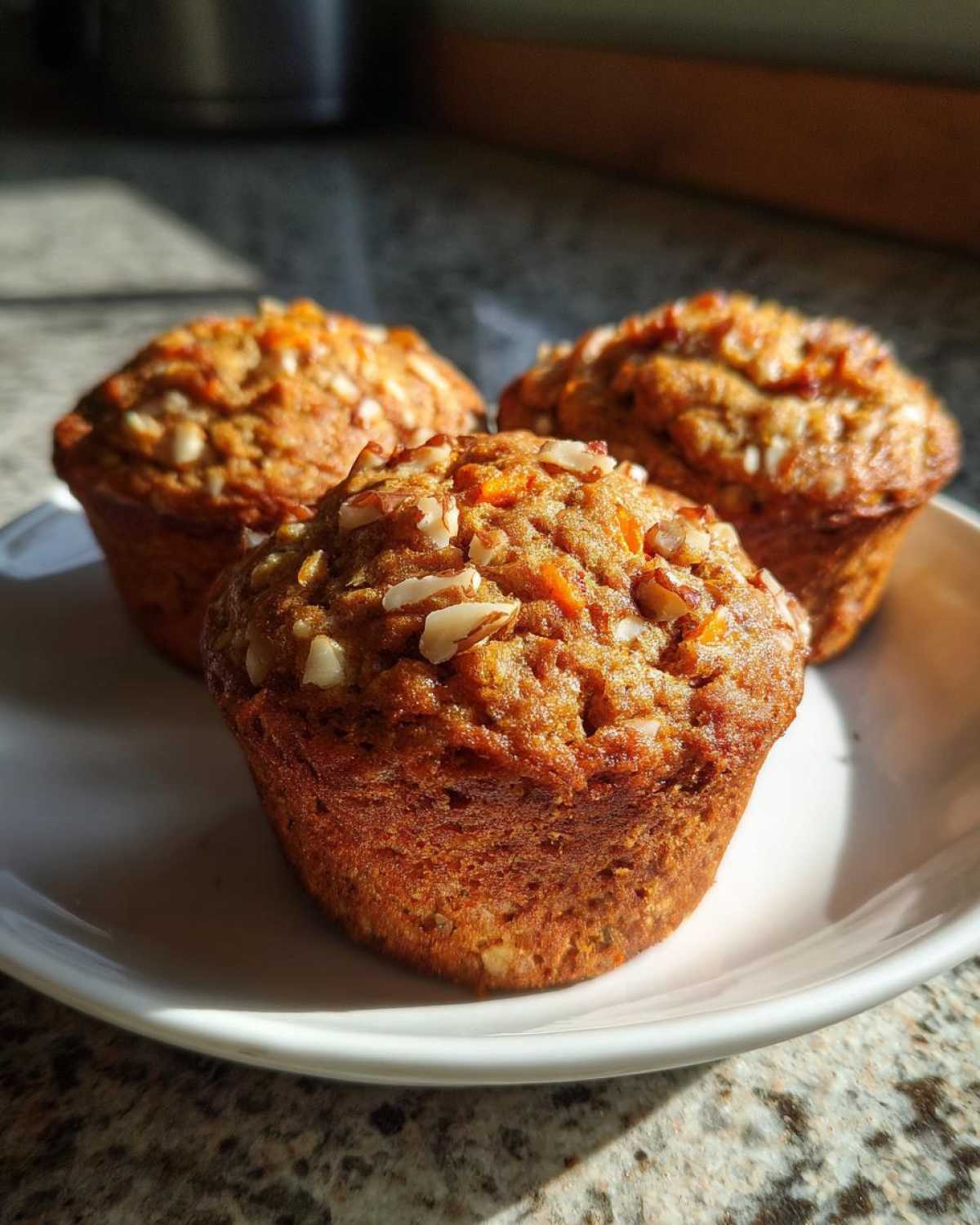 Three freshly baked morning glory muffins topped with chopped nuts, sitting on a white plate in sunlight.