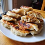 A stack of golden, pan-fried Traditional Chinese Dumplings on a white plate, one cut open showing the savory filling.