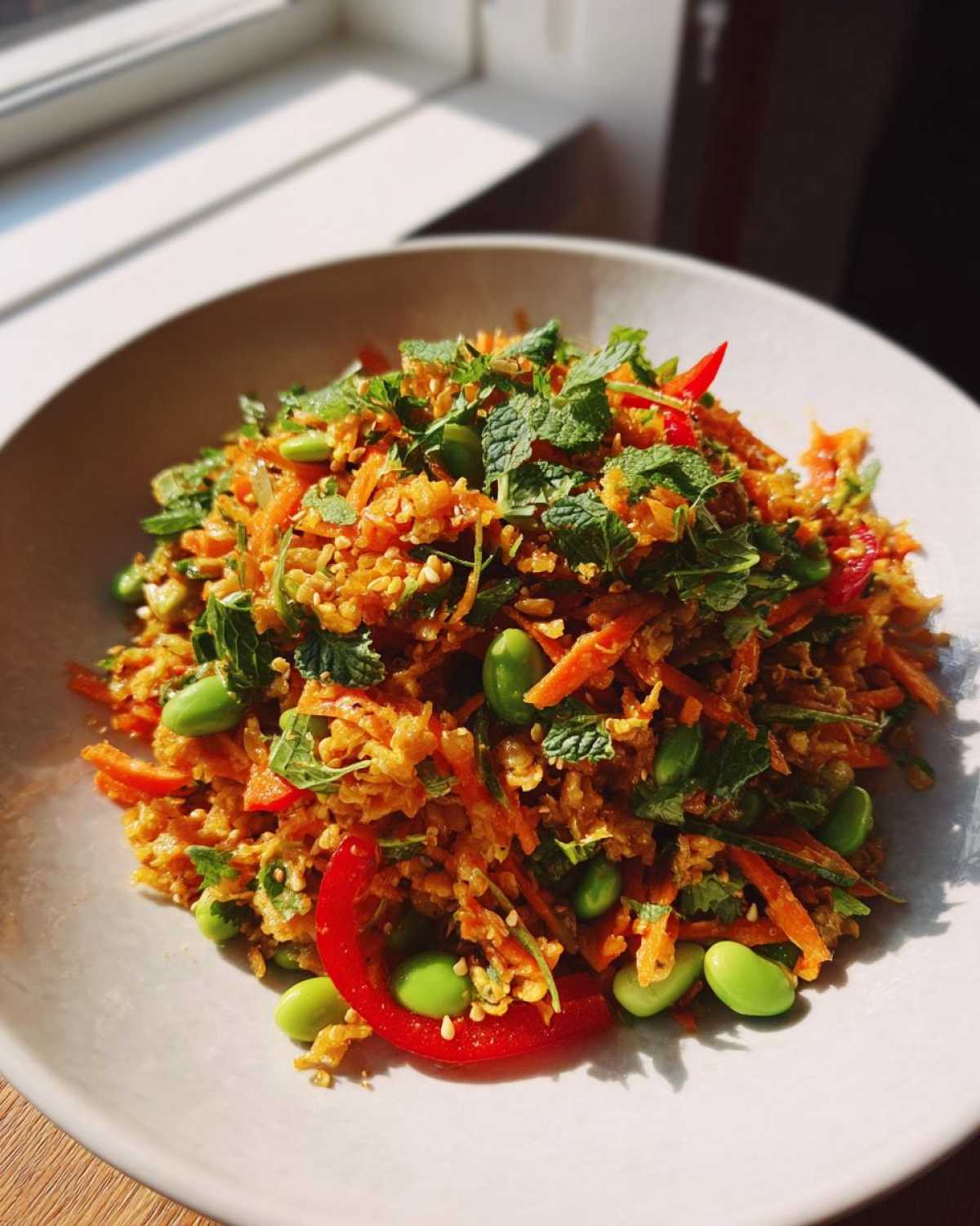 A close-up of a vibrant, orange-hued crispy rice salad mixed with shredded carrots, bright green edamame, and fresh mint leaves.
