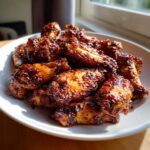 A pile of glistening, caramelized Air Fryer BBQ Chicken Wings served on a white plate, backlit by sunlight.