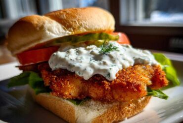Close-up of an Air Fryer Crispy Fish Sandwich featuring a golden fried fillet topped with creamy tartar sauce and dill.