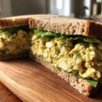 Two halves of an Avocado Egg Salad Sandwich filled with creamy yellow salad and lettuce on whole wheat bread.