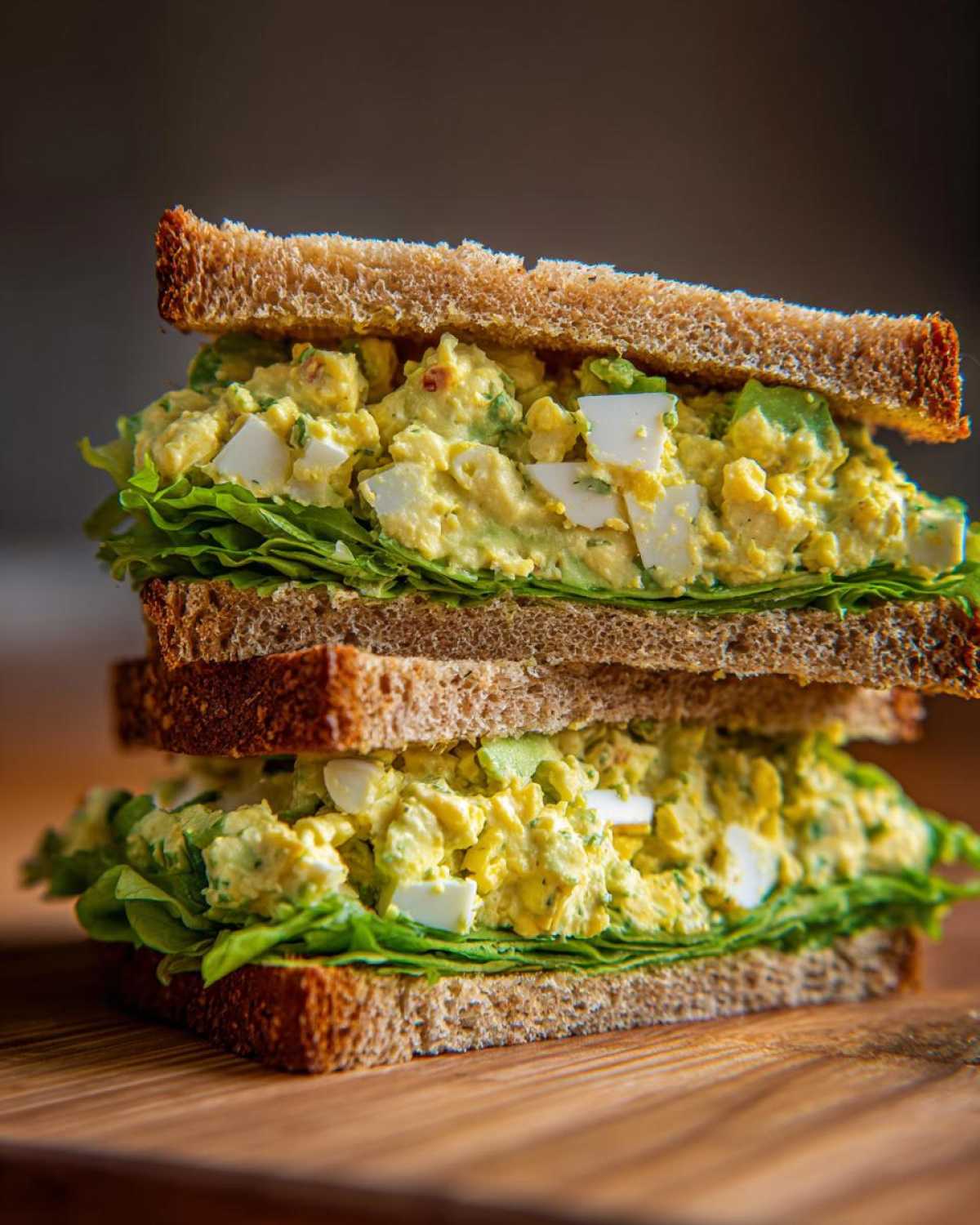 A tall, stacked Avocado Egg Salad Sandwich featuring creamy filling, visible egg chunks, and crisp lettuce on whole wheat bread.