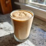 A tall glass filled with a thick, creamy Banana Peanut Butter Smoothie sitting on a sunlit granite countertop.