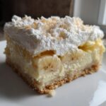 A close-up of a single serving of Banana Pudding Cheesecake Bars showing the crust, creamy filling, banana slices, and whipped topping.