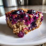 A close-up shot of a moist square of Blueberry Baked Oatmeal Squares topped with jammy blueberries on a white plate.