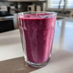 A close-up of a vibrant, deep purple Blueberry Lemon Chia Smoothie in a clear glass on a white kitchen counter.