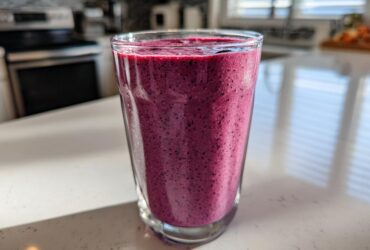 A close-up of a vibrant, deep purple Blueberry Lemon Chia Smoothie in a clear glass on a white kitchen counter.