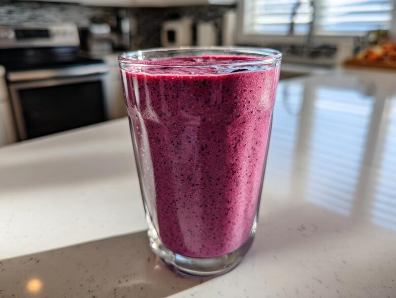 A close-up of a vibrant, deep purple Blueberry Lemon Chia Smoothie in a clear glass on a white kitchen counter.