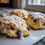 Close-up of three freshly baked Blueberry Lemon Scones topped with a thick white glaze, sitting on a white serving dish.