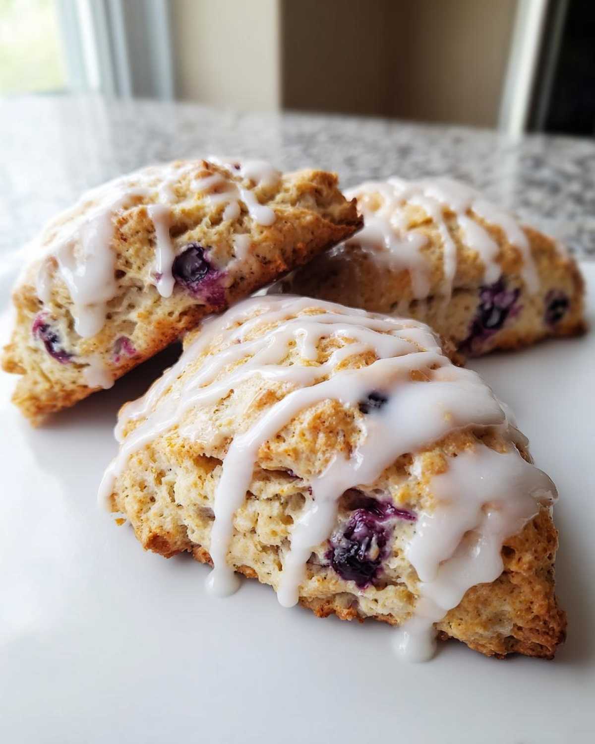 Three freshly baked Blueberry Lemon Scones drizzled with white lemon glaze, sitting on a white plate.