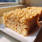 A close-up of thick, golden Brown Butter Rice Krispie Treat Bars resting on a white rectangular plate near a window.