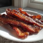 Close-up of crispy, caramelized Brown Sugar Glazed Bacon strips piled on a white plate, glistening in sunlight.