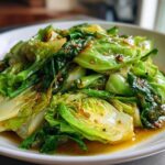 A close-up of vibrant green cabbage stir fry glistening with a savory garlic ginger sauce on a white plate.