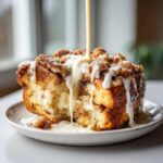 A close-up of a serving of Cinnamon Roll Overnight French Toast Bake being drizzled with thick white icing.
